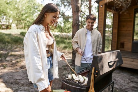 Smiling woman cooking vegetables in grill near blurred boyfriend and vacation house during picnic