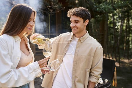 Smiling man pouring wine near girlfriend and blurred bbq with vacation house ay background outdoors