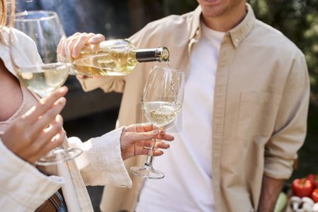 Cropped view of man pouring wine in glasses near blurred girlfriend during picnic outdoors