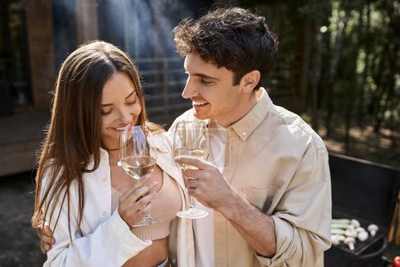 Fröhlicher Mann umarmt Freundin mit Wein in der Nähe von Grill und Ferienhaus im Hintergrund