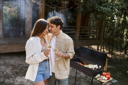 Positive couple holding wine near blurred barbecue and vacation house at background outdoors