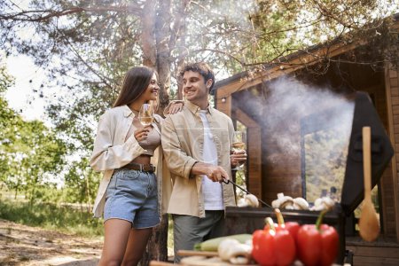 Femme souriante tenant du vin et debout près du petit ami, barbecue avec maison de vacances à l'arrière-plan