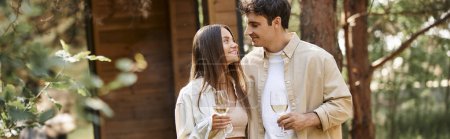 Cheerful love couple with glasses of wine spending time during picnic near vacation house, banner