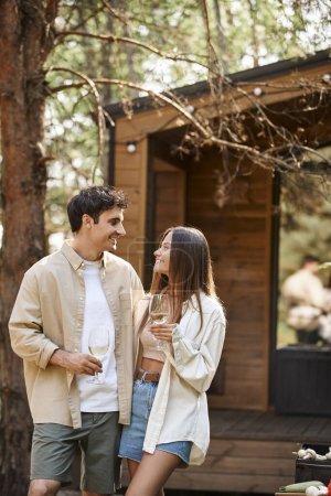 Positive brunette couple holding wine during picnic near barbecue and vacation house at background