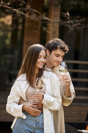 Sonriente hombre sosteniendo el vino y abrazando novia cerca de la parrilla y borrosa casa de vacaciones en el fondo
