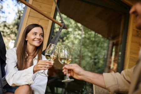Lächelnde Frau stößt mit verschwommenem Freund auf Wein an und sitzt in Hängematte in der Nähe von Ferienhaus