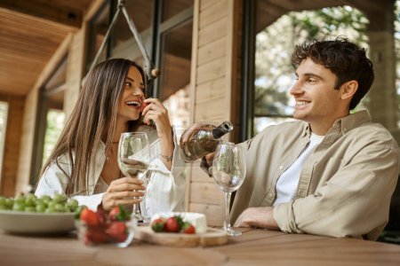 Smiling woman eating strawberry near boyfriend with wine on terrace of wooden vacation house