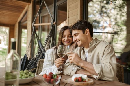 Lächelndes romantisches Paar bei Wein in der Nähe von verschwommenem Essen auf der Terrasse eines hölzernen Ferienhauses