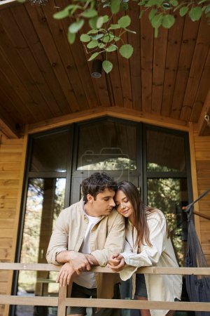 Foto de Morena hombre de pie cerca de novia sonriente con los ojos cerrados en la terraza de la casa de vacaciones de madera - Imagen libre de derechos