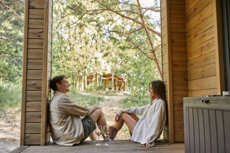 Téléchargez les photos : Vue latérale du couple romantique positif assis près du vin sur le porche de la maison d'été en bois à l'extérieur - en image libre de droit