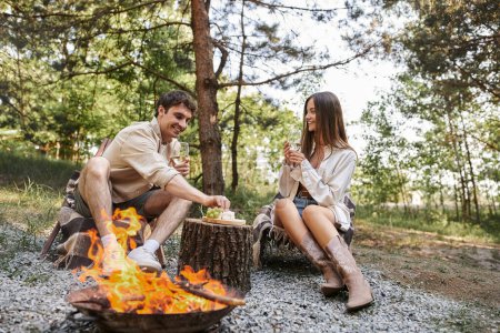 Téléchargez les photos : Couple positif avec du vin et de la nourriture assis sur des chaises longues près du bois de chauffage pendant le pique-nique à l'extérieur - en image libre de droit