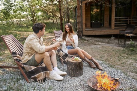 Positive young woman holding wine and talking to boyfriend near food and firewood outdoors