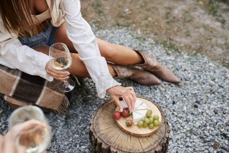 High angle view of young woman holding wine and taking fruits near cheese and boyfriend outdoors