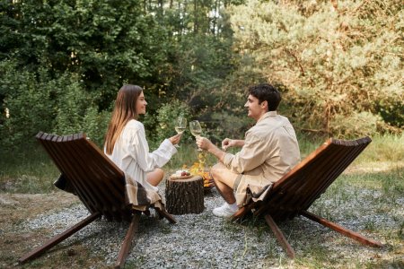 Téléchargez les photos : Vue latérale du couple romantique souriant tenant du vin près de la nourriture et du bois de chauffage pendant le pique-nique à l'extérieur - en image libre de droit