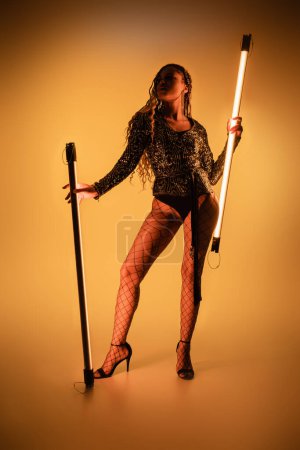 sexy woman and fashion, african american model holding fluorescent lamps on yellow background