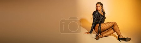 Photo for Banner, dark skinned woman with tattoo sitting on yellow background, fishnet tights and high heels - Royalty Free Image