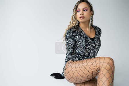 fashion and accessories, dark skinned woman posing in fishnet tights, sitting on grey background
