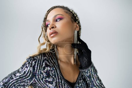 Téléchargez les photos : Portrait de femme afro-américaine en gant ajustant boucle d'oreille, fond gris, mode, détourner les yeux - en image libre de droit