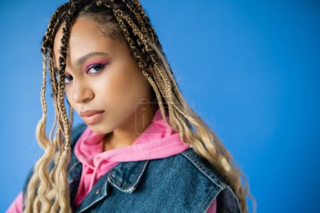 young african american woman with dreadlocks looking at camera on blue background, bold makeup
