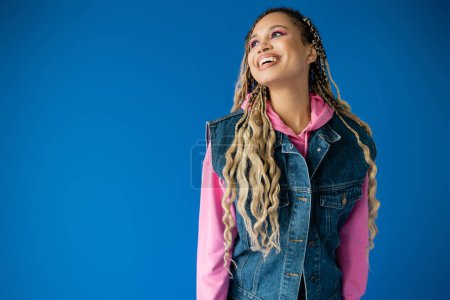 happy african american woman in pink hoodie and denim vest smiling on blue background, bold makeup
