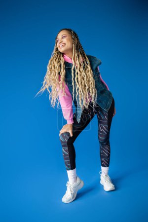 full length, happy african american woman in pink hoodie and denim vest posing on blue background