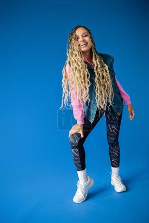 Photo for Full length, joyful african american woman in pink hoodie and denim vest posing on blue background - Royalty Free Image