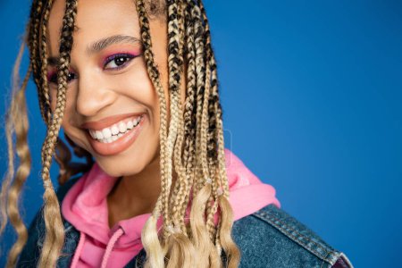 attractive african american woman in pink hoodie and denim vest looking at camera, blue background
