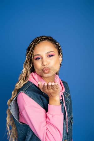 beautiful african american woman sending air kiss on blue background, denim vest and pink hoodie