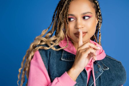 Photo for Attractive african american woman showing shh and looking away, finger near lips, blue background - Royalty Free Image