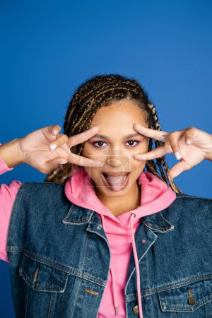 african american woman with dreadlocks showing v sign with hands and sticking out tongue on blue