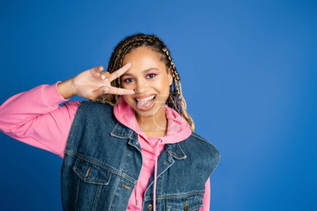 heureuse femme afro-américaine avec dreadlocks montrant le signe v avec la main, sortant la langue sur bleu