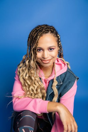 Photo for Dark skinned woman in pink hoodie, denim vest and leggings looking at camera on blue background - Royalty Free Image