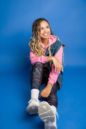 happy african american woman with dreadlocks sitting on blue background and looking away, sporty