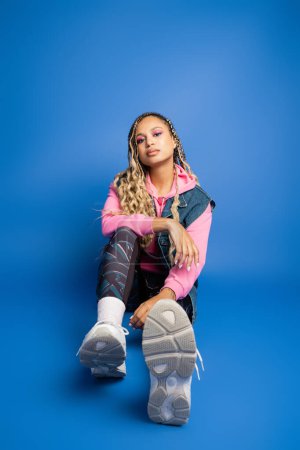 confident african american woman with dreadlocks sitting in sporty outfit on blue background