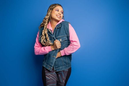 Photo for Happy woman in denim vest and pink hoodie standing on blue background, looking away in studio - Royalty Free Image