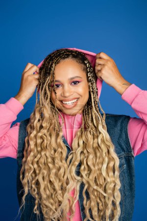 positive dark skinned woman wearing pink hood on head, looking at camera on blue background