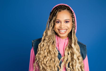 african american woman with bold makeup wearing hoodie and denim vest, smiling in studio on blue