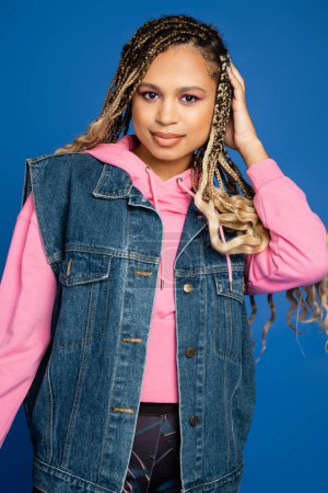 Photo for Dark skinned woman with bold makeup smiling on blue, wearing pink hoodie and denim vest in studio - Royalty Free Image