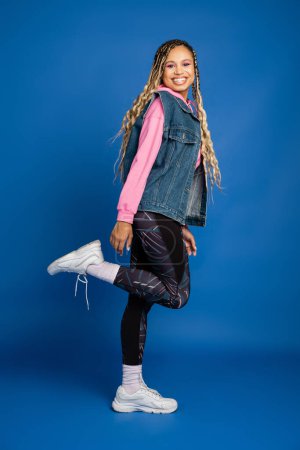 dark skinned woman in sporty outfit smiling on blue background, positive model with bold makeup