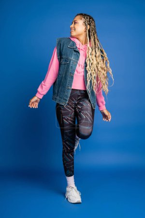 full length, dark skinned woman in sporty outfit smiling on blue background, positive, bold makeup