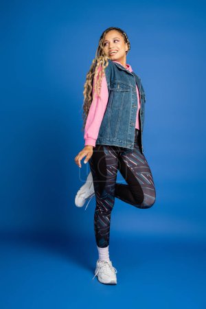 full length, dark skinned woman in sporty outfit posing on blue background, positive, bold makeup