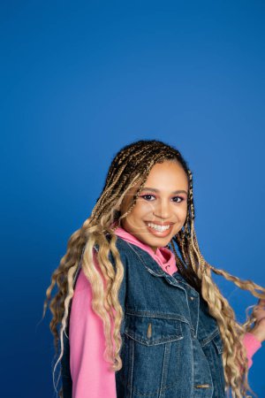 Photo for Portrait, happy dark skinned woman standing on blue background, denim vest and pink hoodie, smile - Royalty Free Image