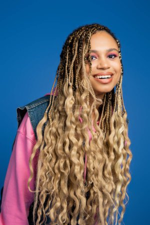 charming african american woman with long hair smiling and looking at camera on blue background