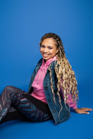 happy african american woman with long hair looking at camera, sitting on blue background, sporty