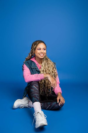 longitud completa de la joven afroamericana de pelo largo sentada en traje deportivo sobre fondo azul