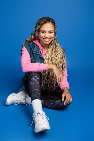 full length of happy long haired african american woman sitting in sporty outfit on blue background
