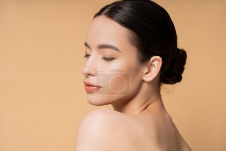 Beautiful young asian woman with closed eyes and naked shoulders standing isolated on beige