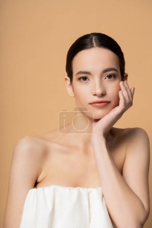 Young brunette asian model in top with naked shoulders looking at camera isolated on beige