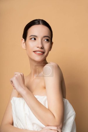 Foto de Sonriente joven mujer asiática en la parte superior con los hombros desnudos de pie aislado en beige - Imagen libre de derechos
