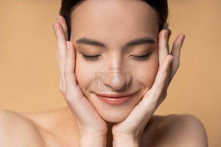 Close up view of asian woman with closed eyes and naked shoulders touching face isolated on beige